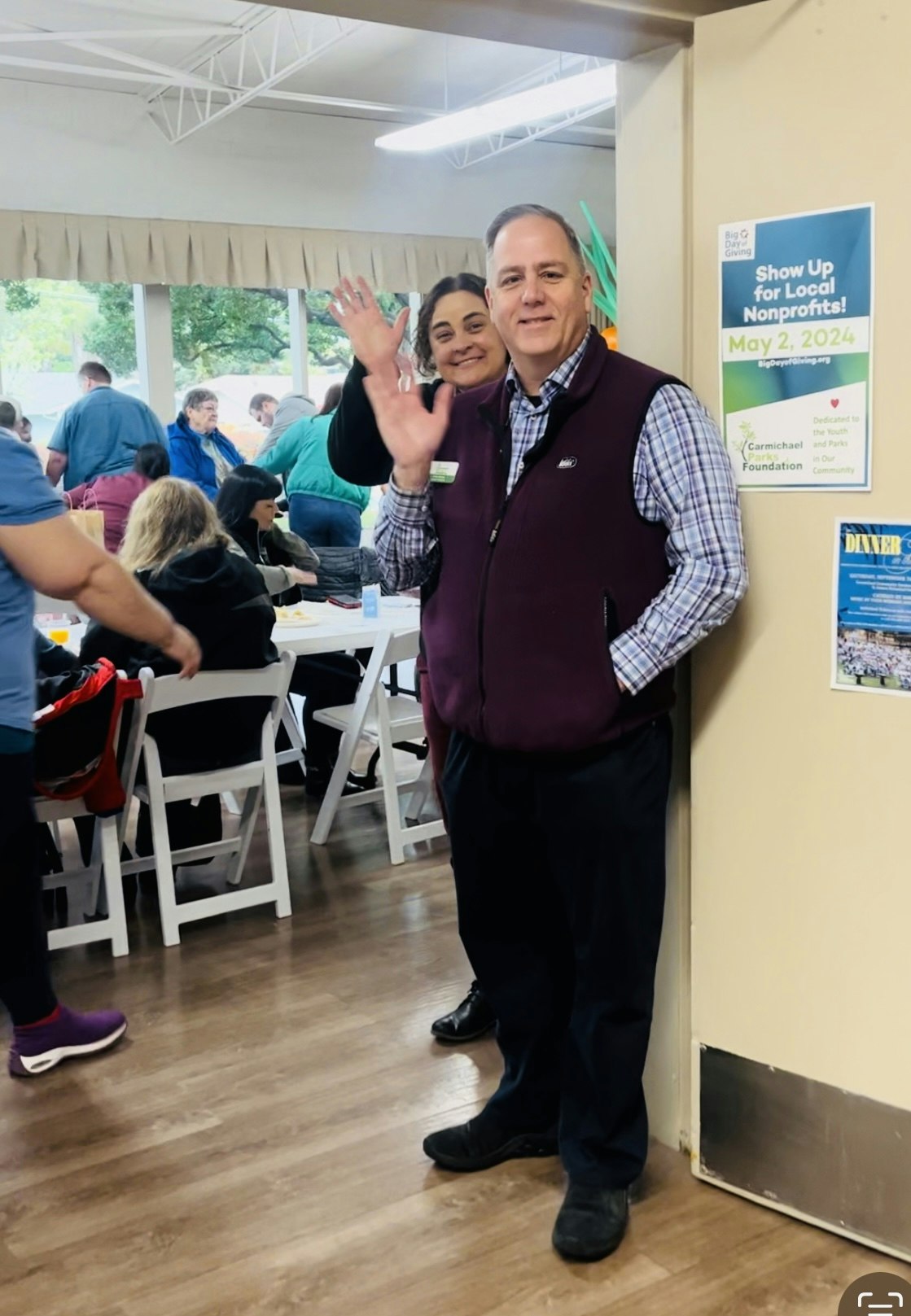 Two people smiling and waving in a busy room with tables and a poster about local nonprofits.