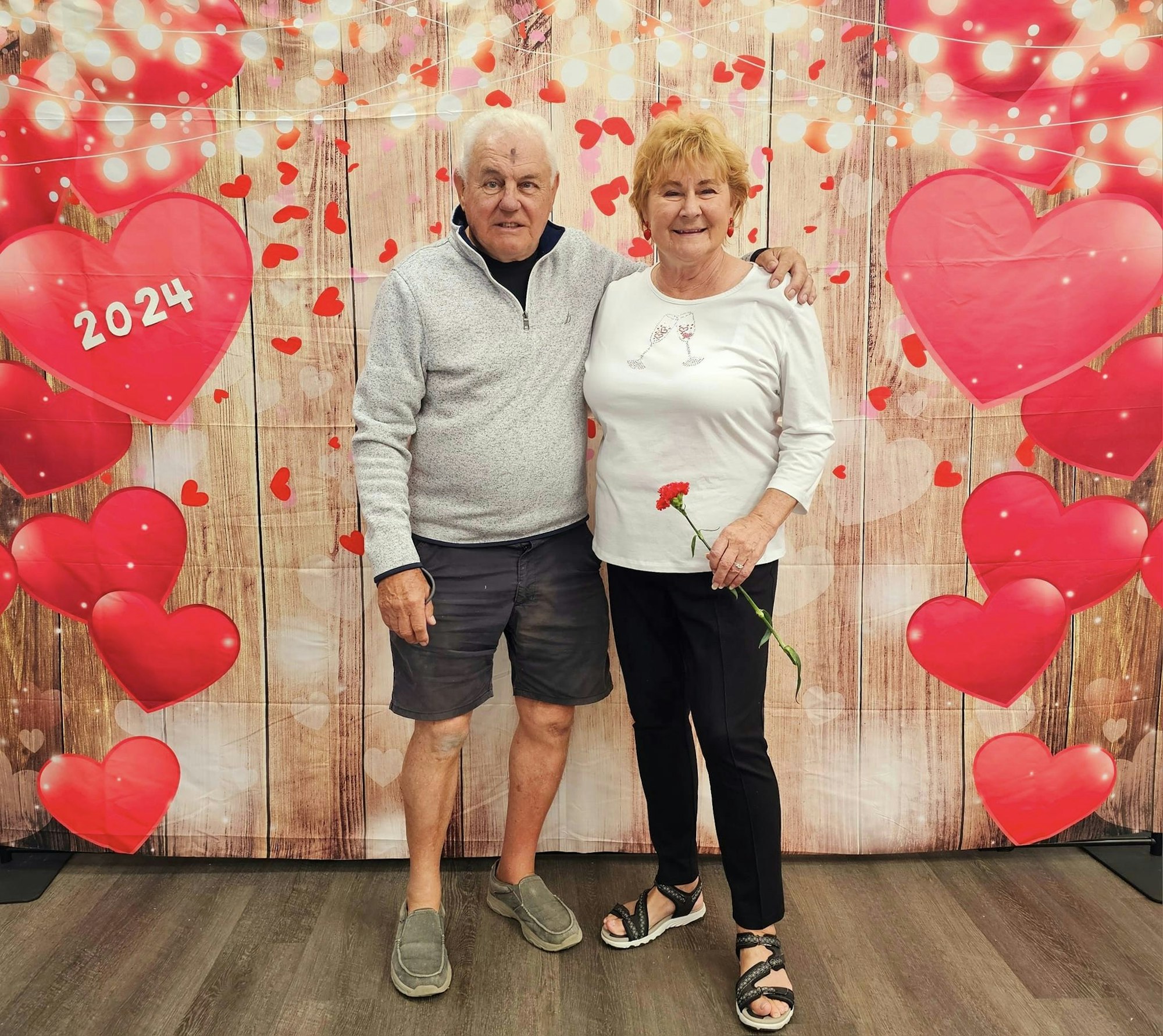 Two people posing with a Valentine's Day themed backdrop, heart decorations, and a date "2024".