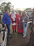 Four people pose together outdoors, with one dressed as Santa Claus, near vehicles, during what appears to be a festive gathering.