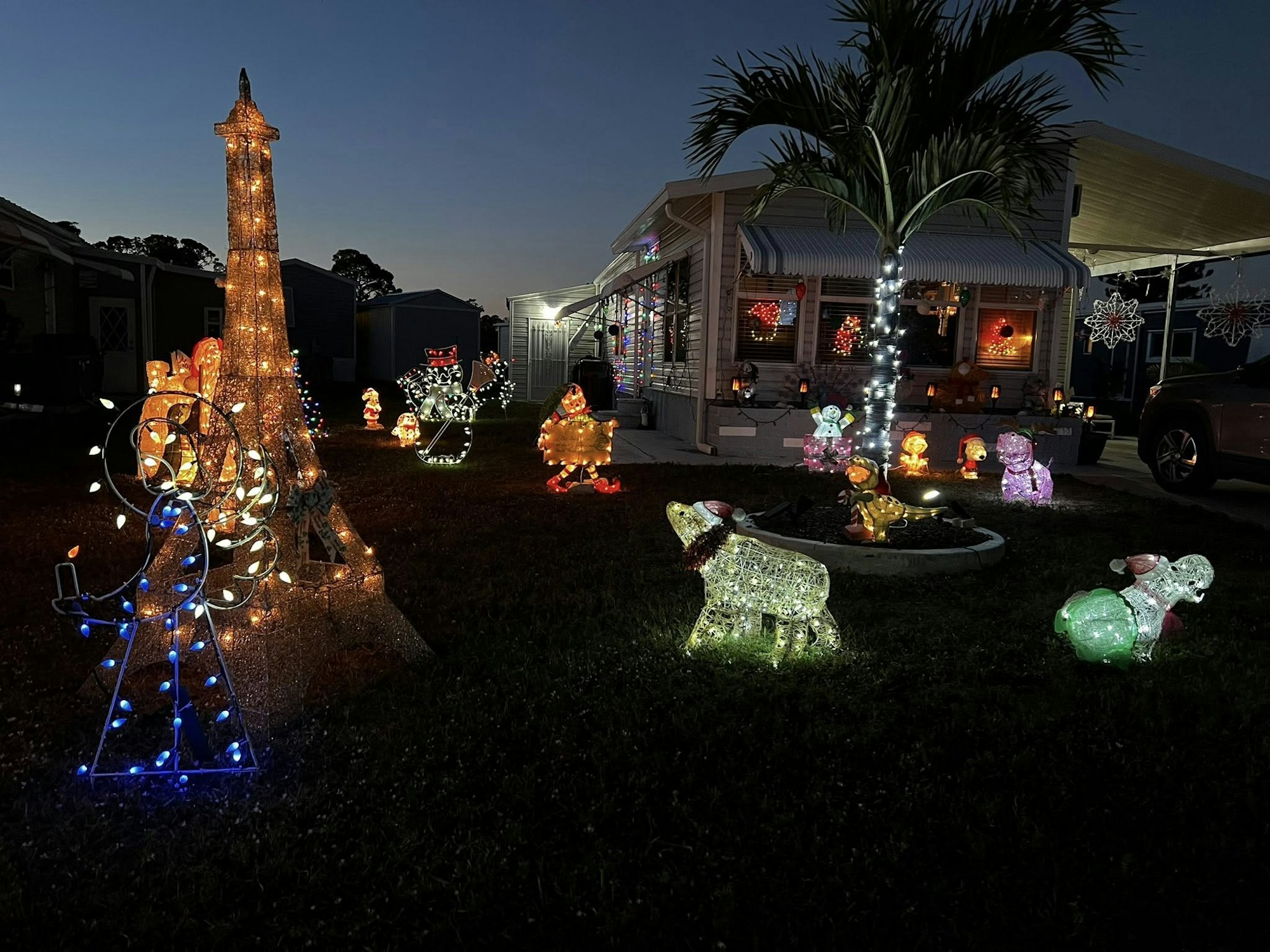 A festive display of holiday lights, featuring a mini Eiffel Tower, snowmen, animals, and cheerful decorations in a yard at dusk.