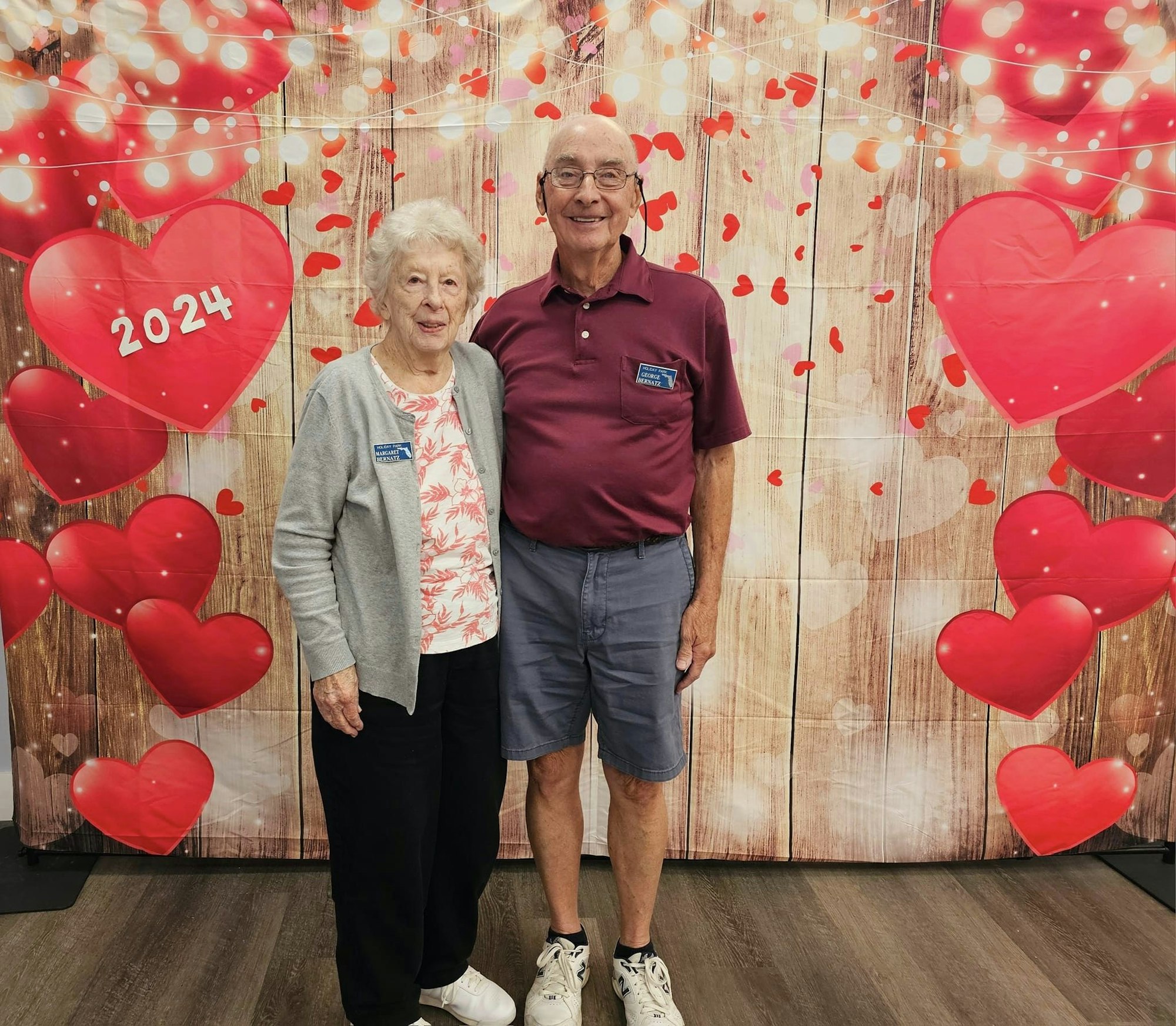 An elderly couple smiles in front of a decorative backdrop with hearts and lights, celebrating a themed event for 2024.