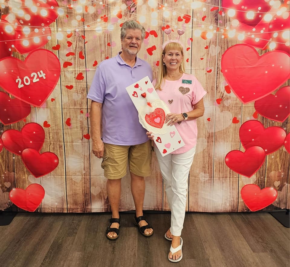 A couple stands smiling, holding a large heart-shaped card, with a backdrop of hearts and "2024" displayed, in a Valentine's-themed setting.