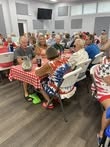 A group of people sitting at tables with red and white checkered tablecloths, engaged in conversation in a bright room.