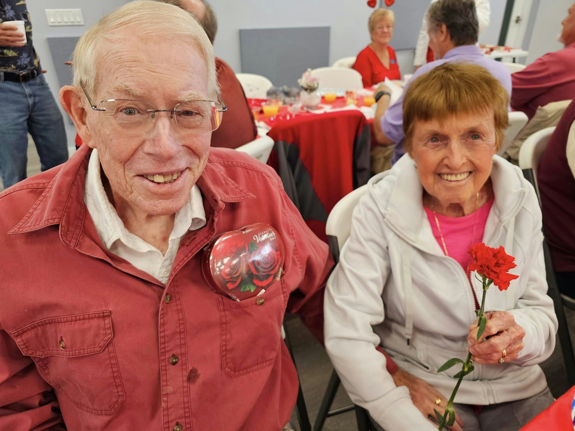 Elderly couple smiling, man with Valentine's balloon, woman holding a red flower, festively decorated venue.