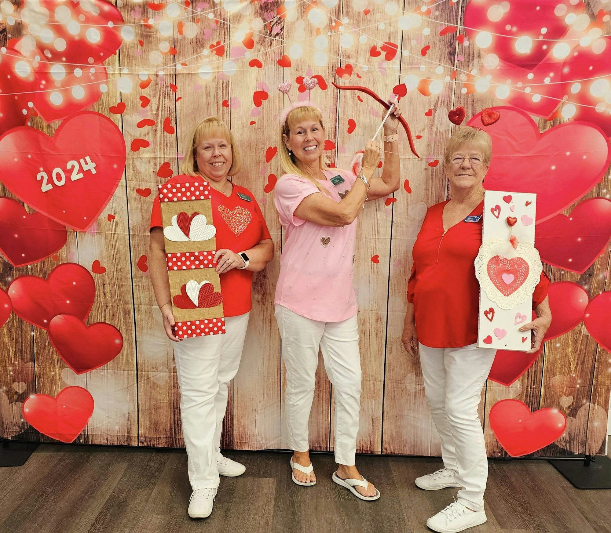 Three people in festive attire posing with heart decorations in a Valentine's Day-themed setup.