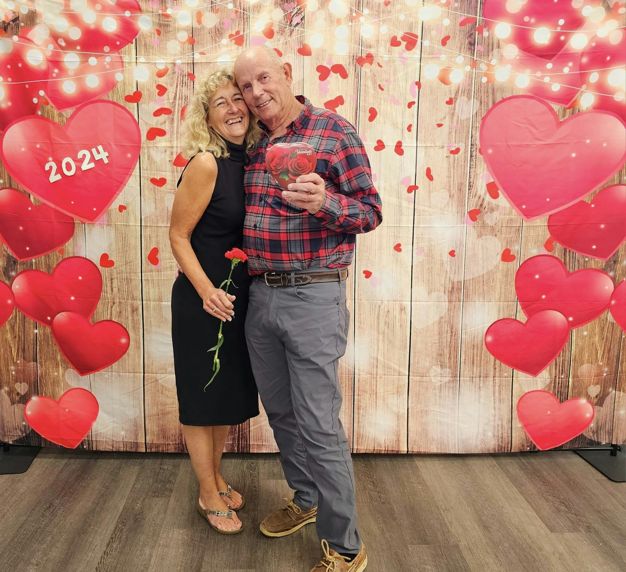 A smiling couple posing with a rose and a heart-shaped box against a backdrop with hearts and "2024" text. #ValentinesDayVibes