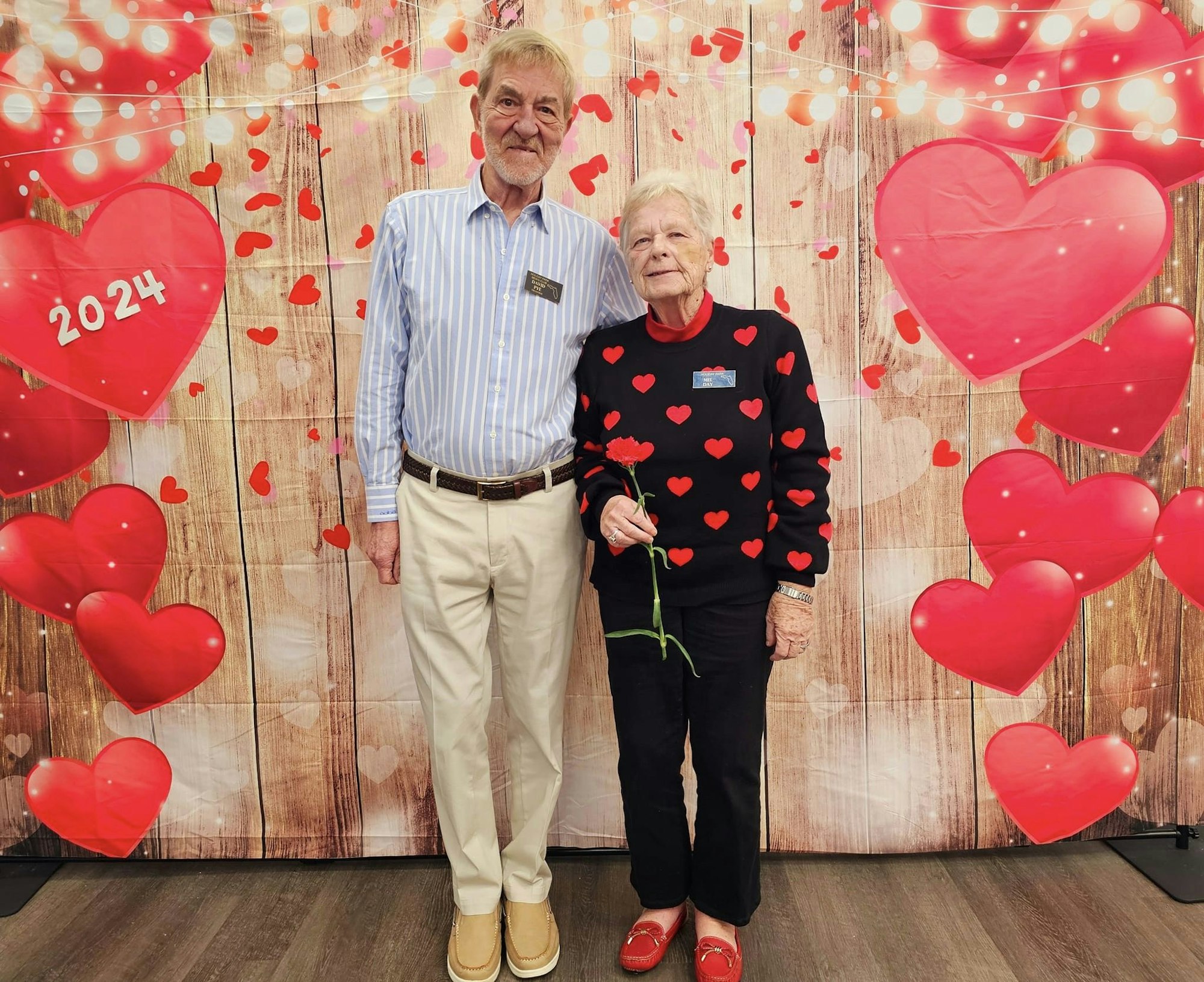 Two seniors stand before a heart-themed backdrop with a "2024" heart, likely celebrating Valentine's Day or a similar event.