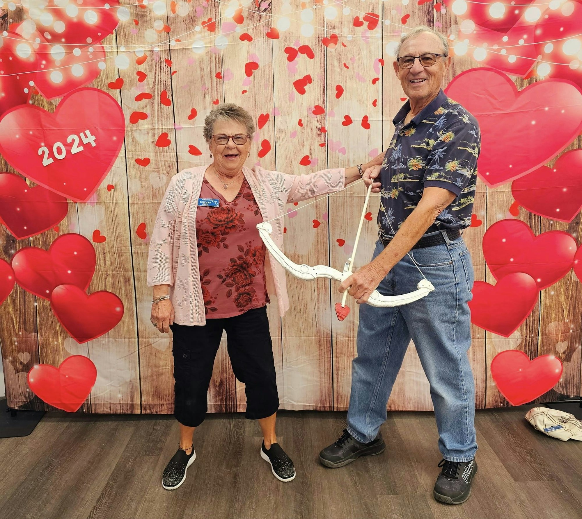 Two people with a heart-themed backdrop holding a large decorative bow and arrow, smiling. #ValentinesDay