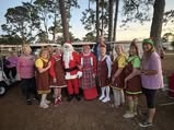 A festive group featuring Santa and several people in themed outfits, posing together outdoors among trees.