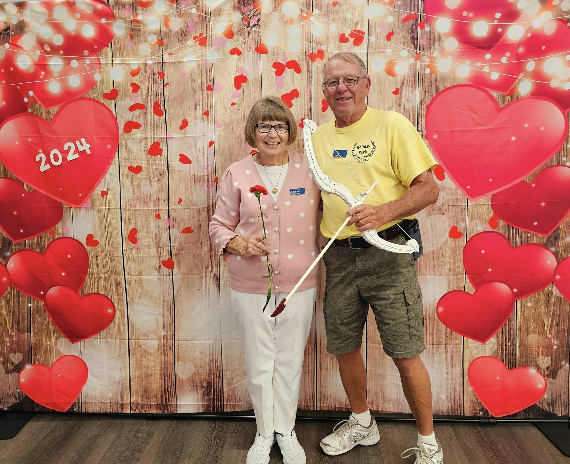 An older couple with a rose and crutch, standing in a room decorated with heart balloons and a "2024" heart sign.