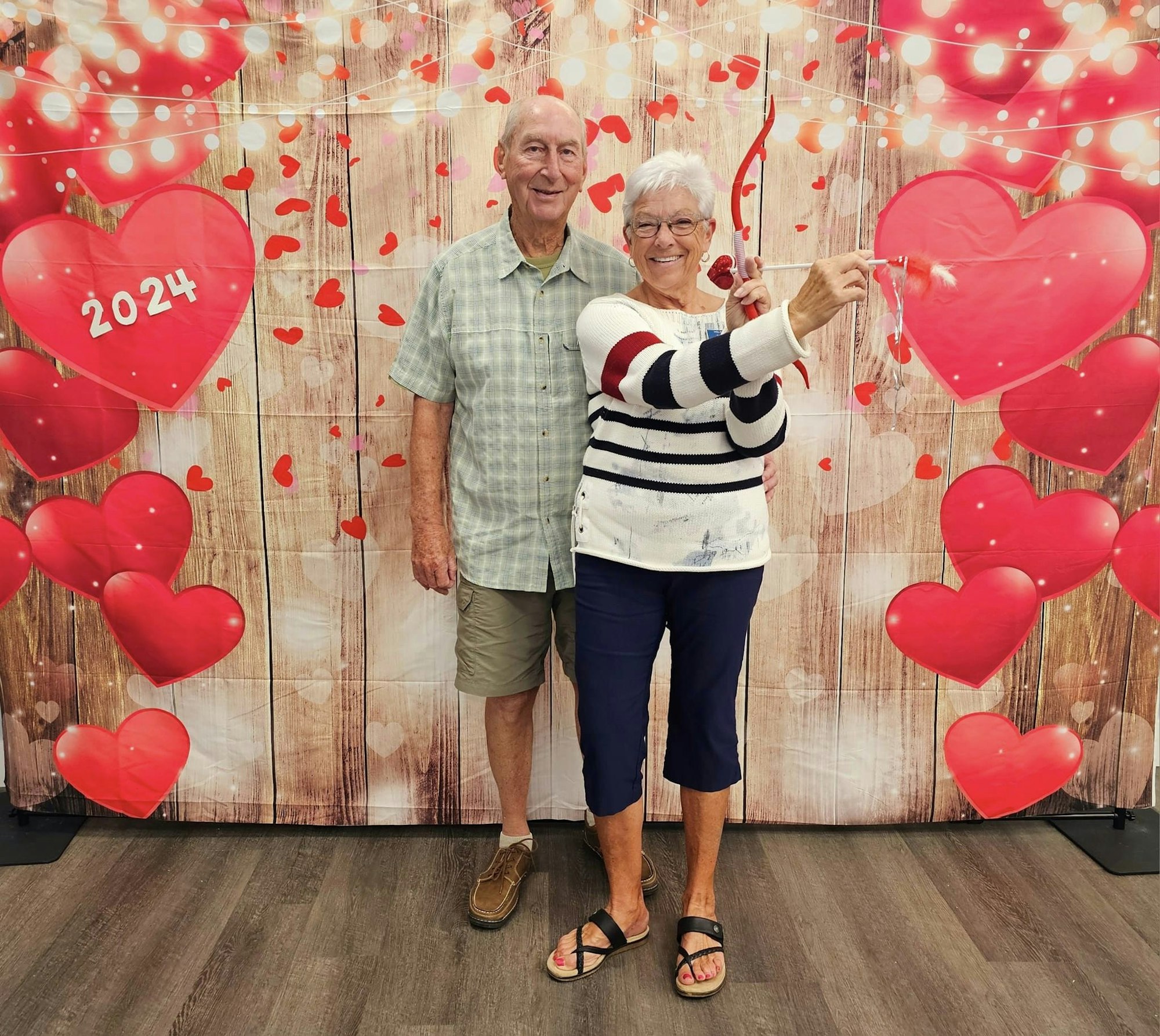 An elderly couple posing together with a festive backdrop of hearts and "2024" for a celebration.