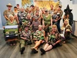 A group of people in camouflage outfits poses together, smiling in a lively setting with a decorative background.