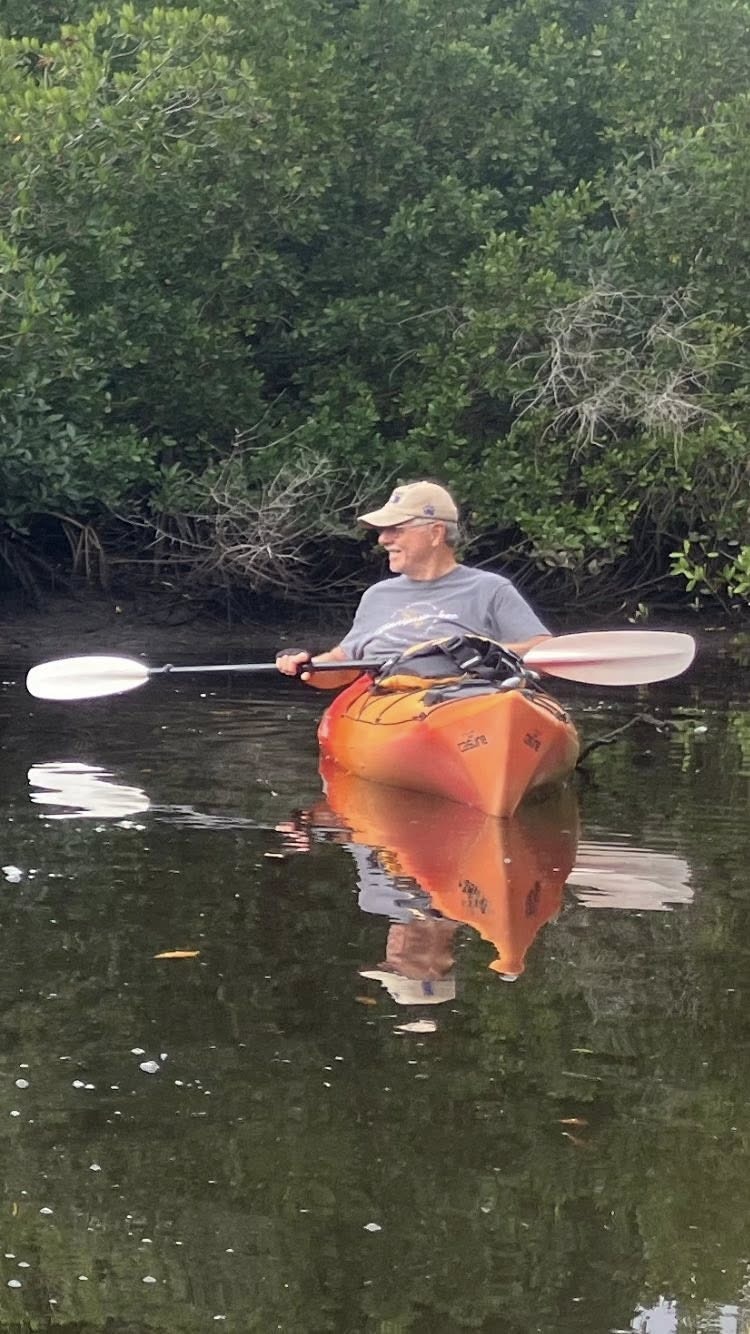 A person is kayaking in calm water, surrounded by lush greenery. The scene reflects tranquility and nature.