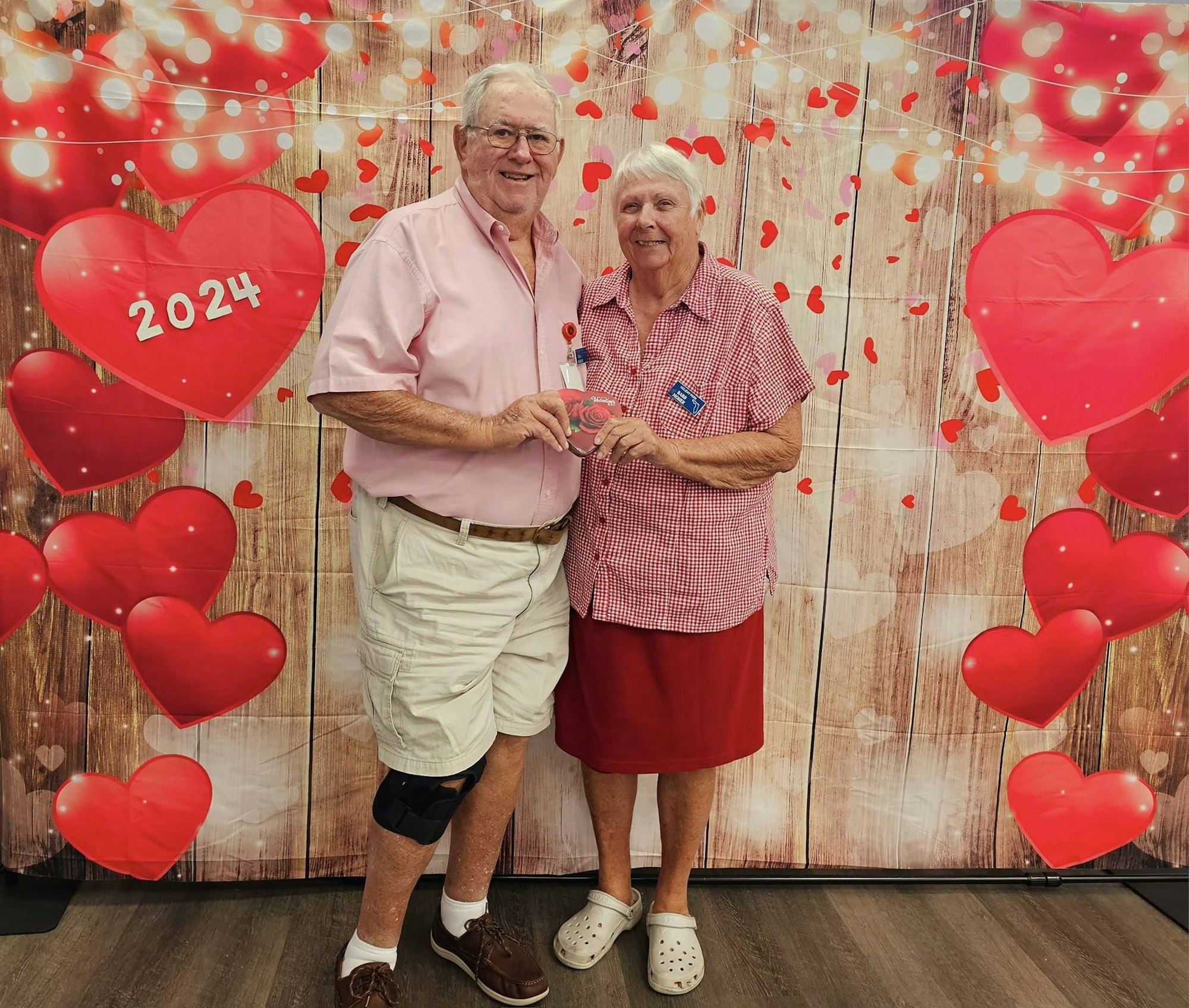 An elderly couple standing together in front of a Valentine’s-themed backdrop with hearts and "2024" written on it.