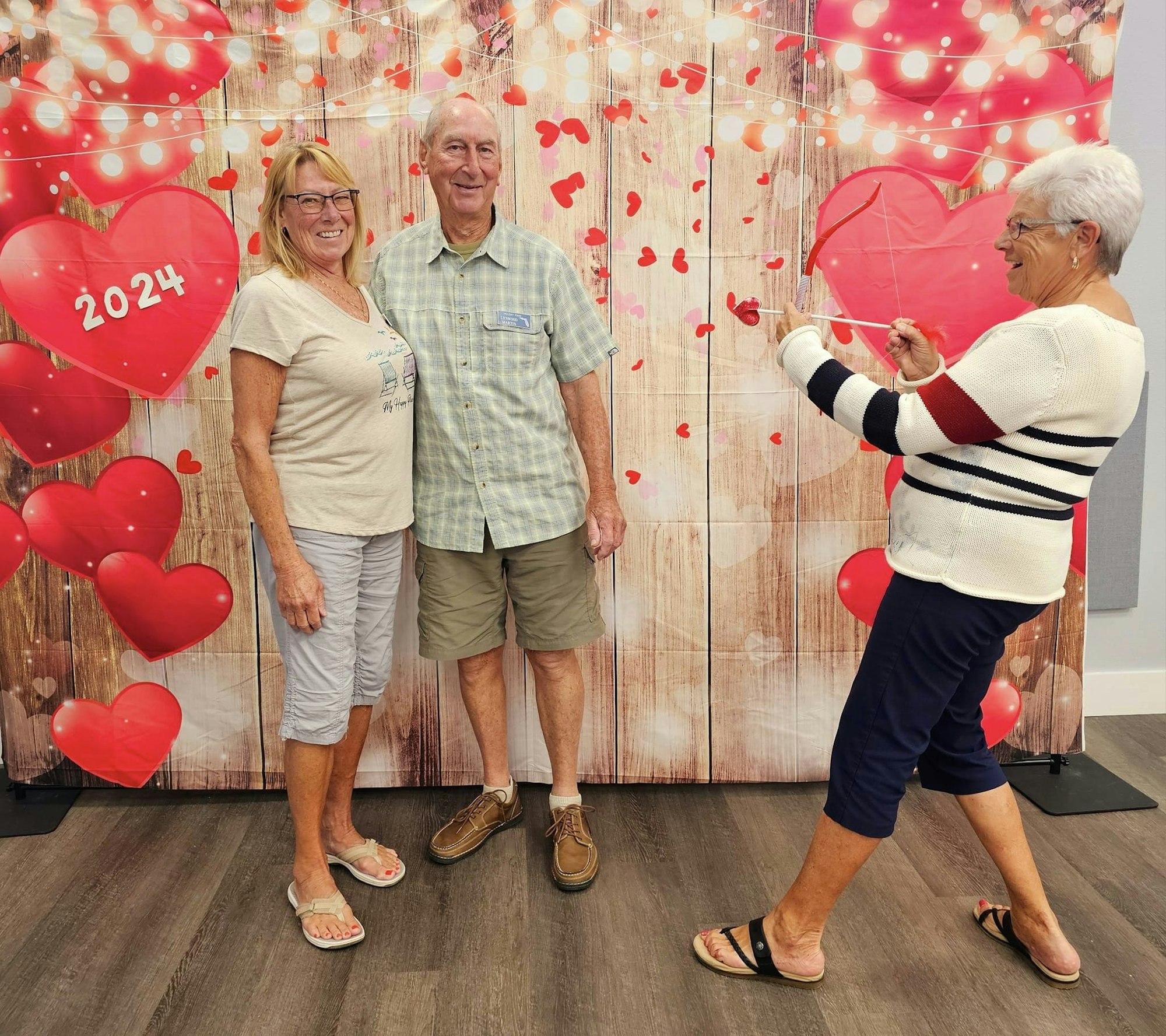 Three seniors posing, two smiling at the camera, one pretending to draw a heart, in front of a Valentine's themed backdrop.