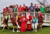 A festive group of people in colorful outfits, some dressed as Santa and holiday characters, posing cheerfully outdoors.
