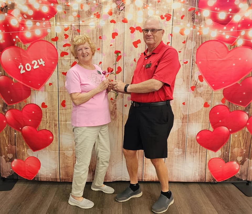 Two people holding hands with a rose, standing in front of a Valentine-themed backdrop with hearts and "2024".