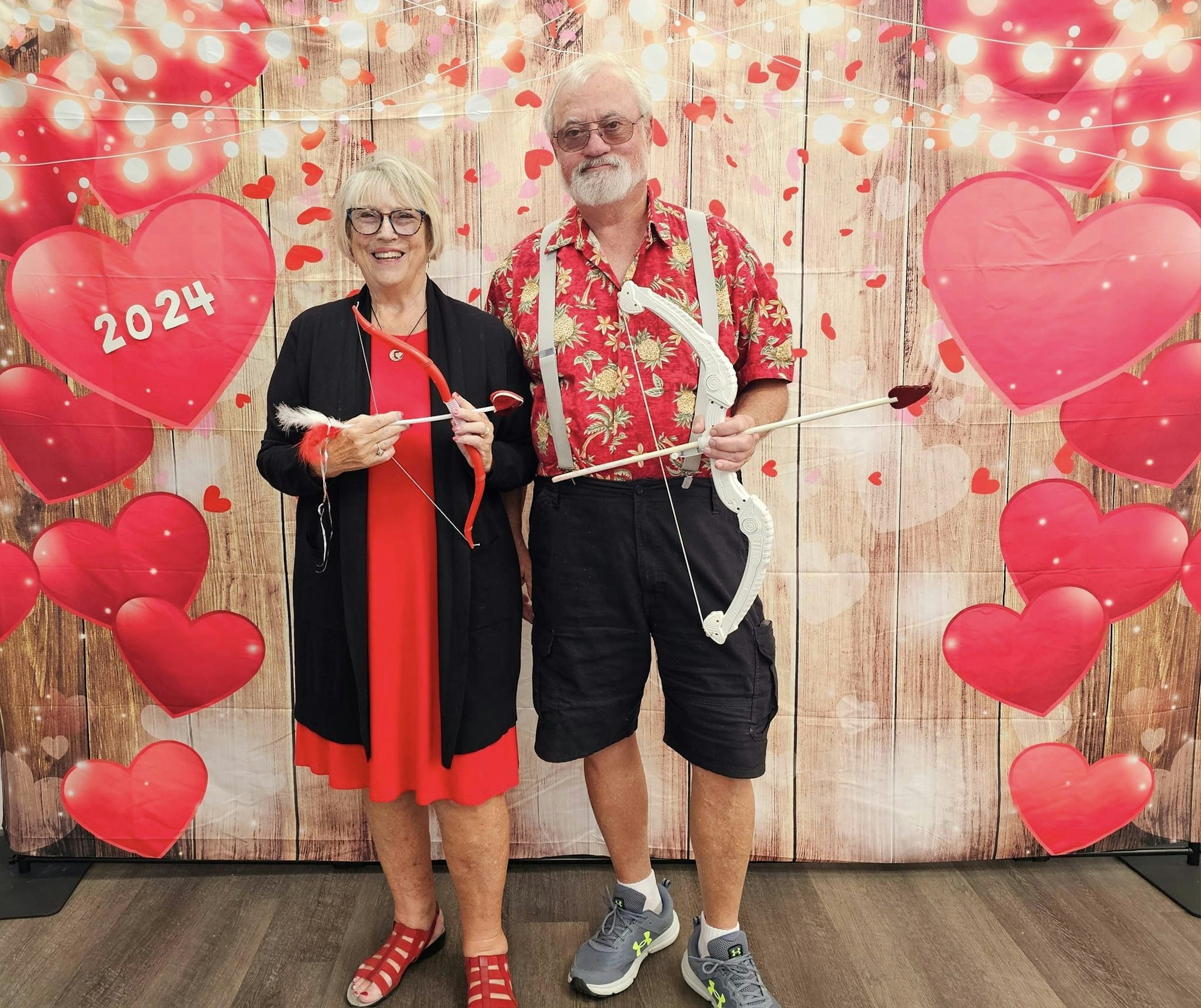 A couple with a cupid's bow and arrow, standing in front of a Valentine-themed backdrop with hearts and "2024" sign.