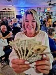 A smiling woman holds a fan of cash, surrounded by a lively crowd in a social setting, possibly a bar or restaurant.