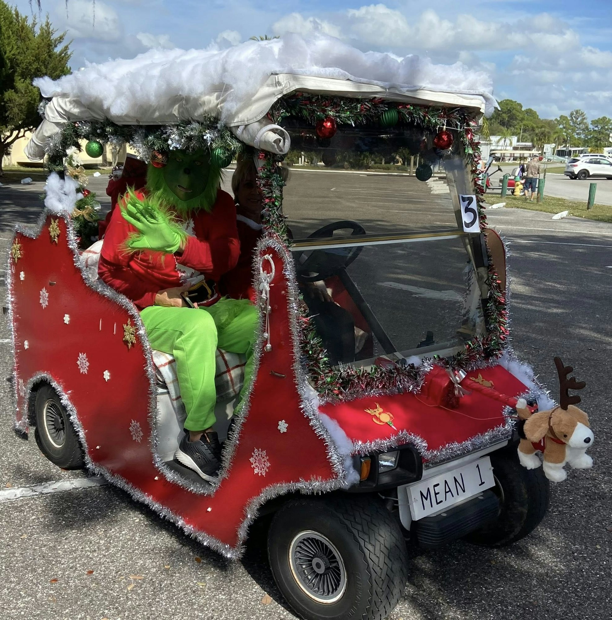 A festive golf cart decorated for Christmas, featuring a person dressed as the Grinch inside. It's adorned with tinsel and ornaments.