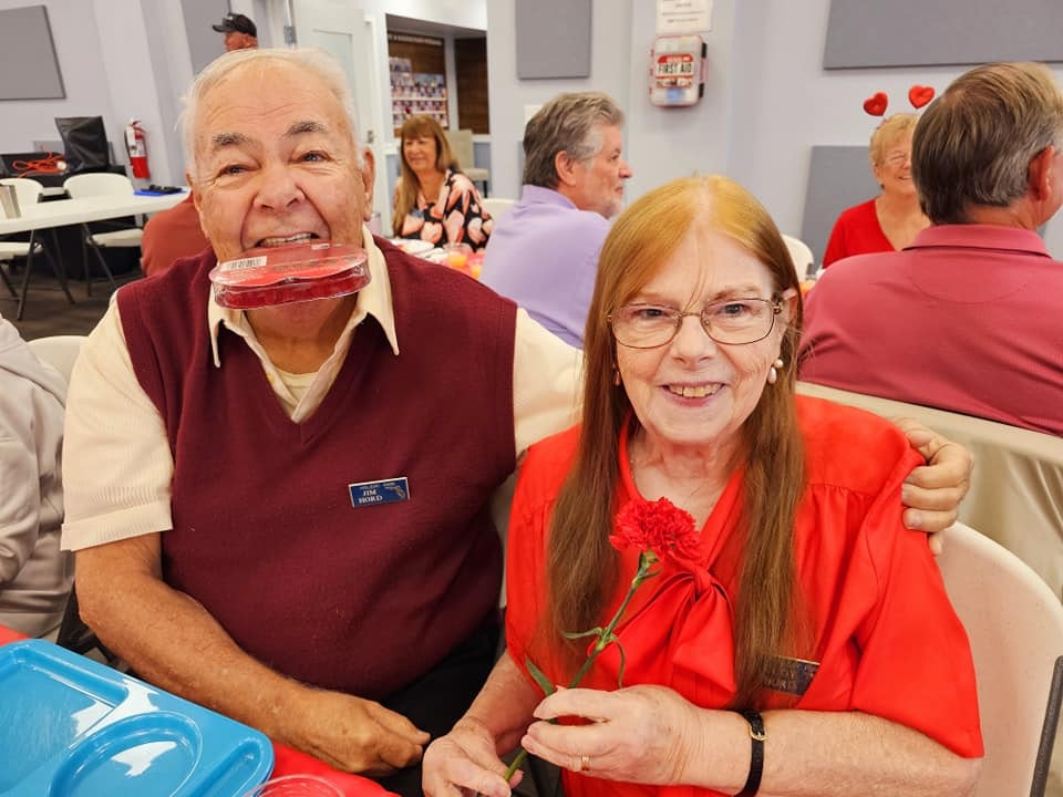 An elderly couple at a social event, man with a dessert on his mouth, woman holding a rose, both smiling.