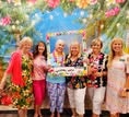 A cheerful group of women in colorful outfits posing together against a vibrant, tropical-themed backdrop.