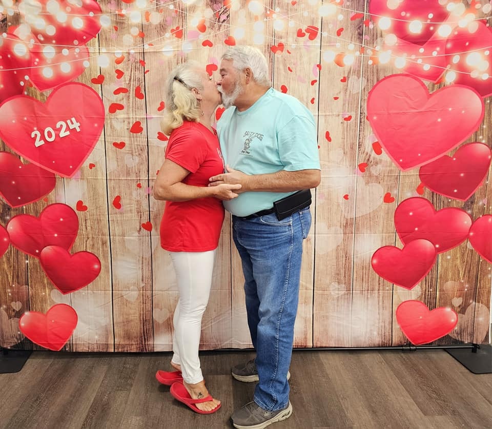 Two people embracing in front of a heart-themed backdrop with "2024" written on it.