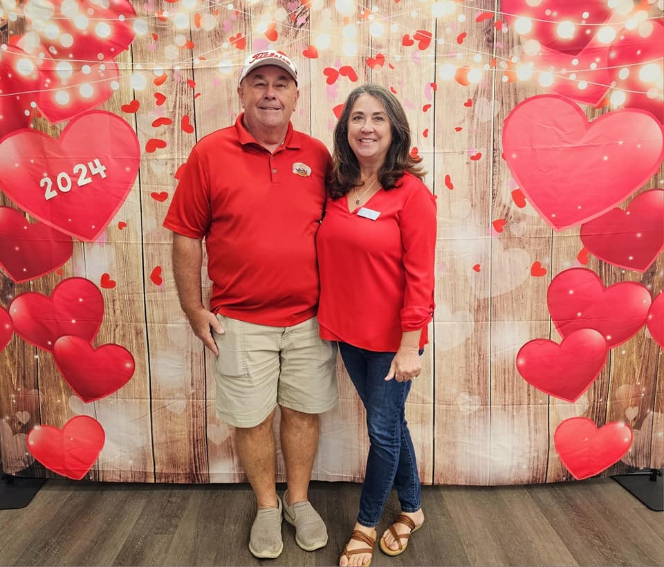A couple stands smiling in front of a Valentine-themed backdrop with hearts and "2024" displayed.