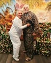 A couple poses together, with one wearing camouflage and the other in white attire, against a tropical-themed background.