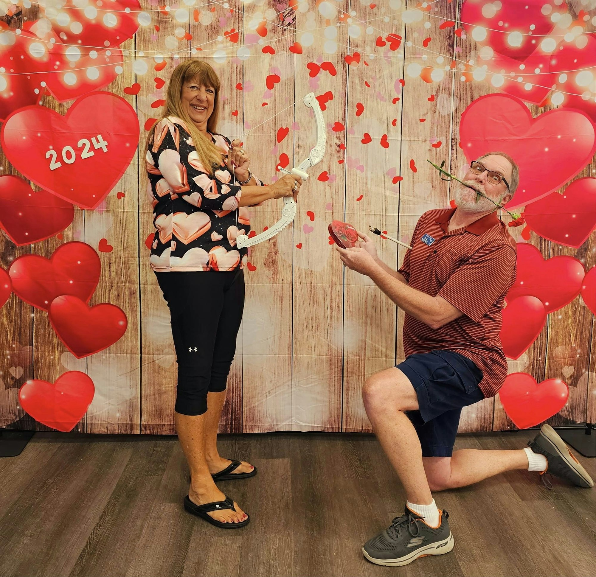 Two people staging a Valentine's Day pose with hearts and Cupid's bow against a festive backdrop.