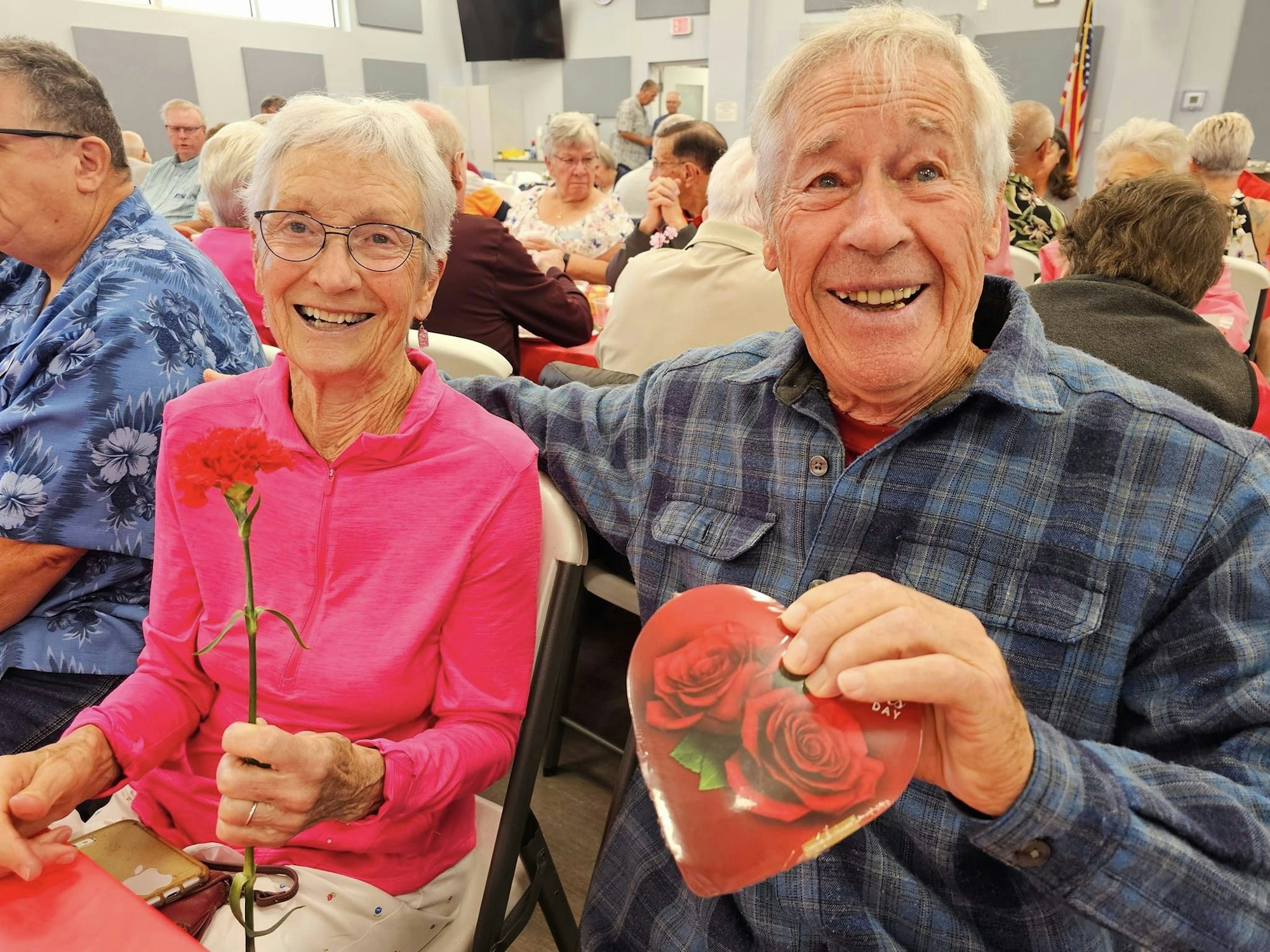 Two happy seniors with a red rose and heart-shaped box, likely celebrating a joyful occasion together.