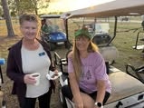 Two women are smiling and posing beside a golf cart, enjoying a casual moment outdoors.