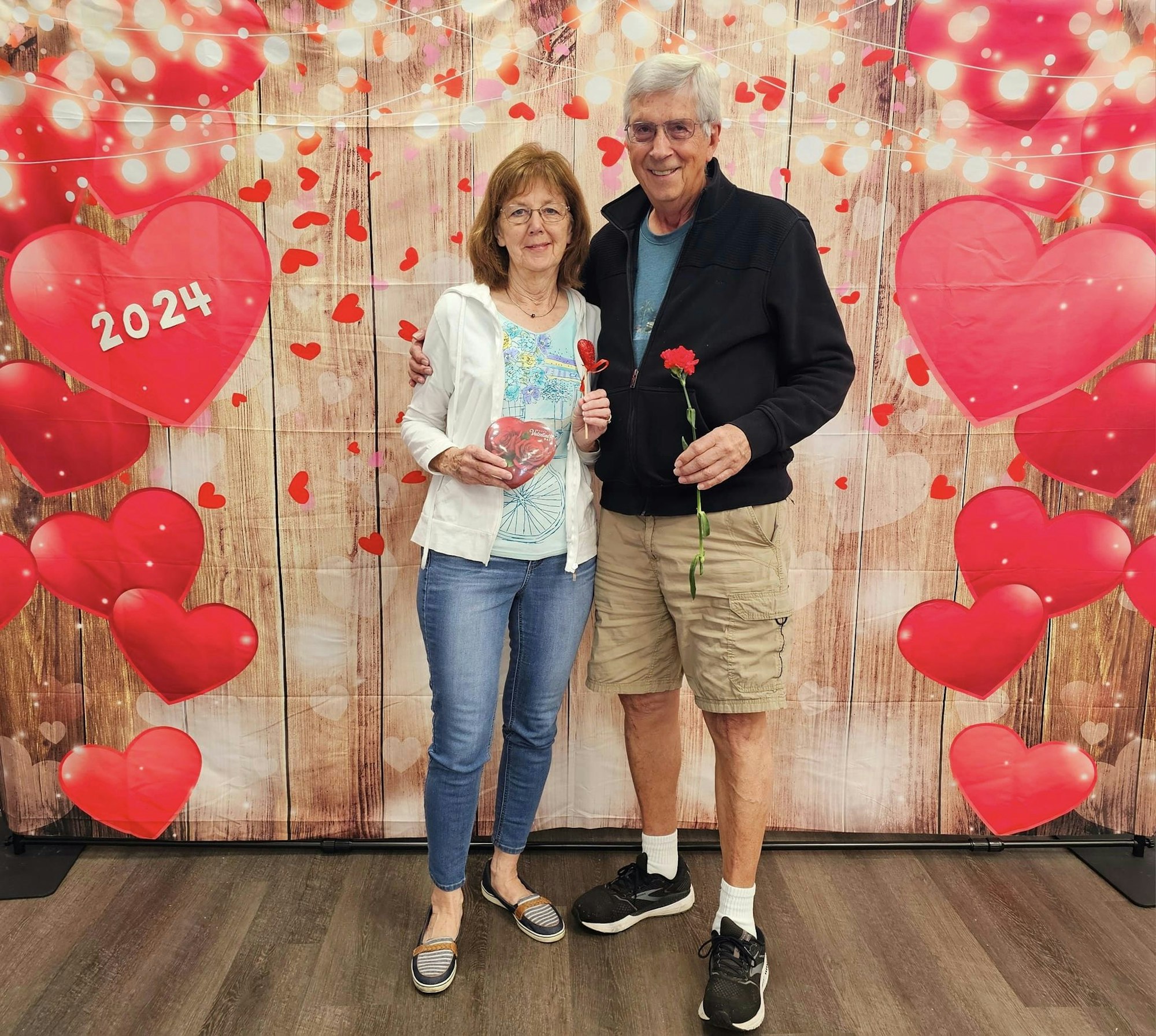 Two people posing with flowers in front of a heart-themed backdrop, likely celebrating Valentine's Day or a romantic occasion.