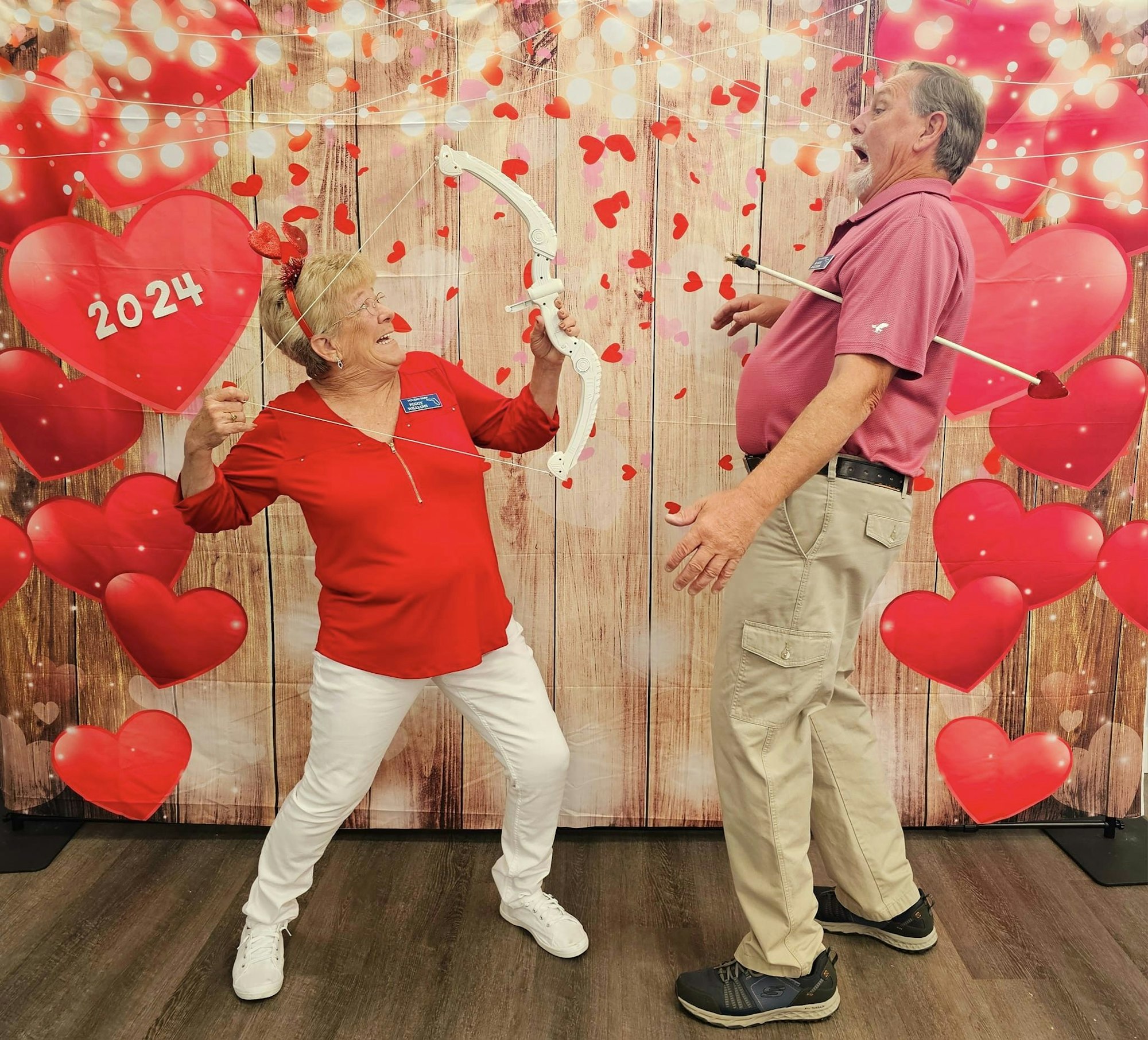 A playful scene with a woman in red pretending to shoot arrows at a surprised man, surrounded by hearts and festive decorations.