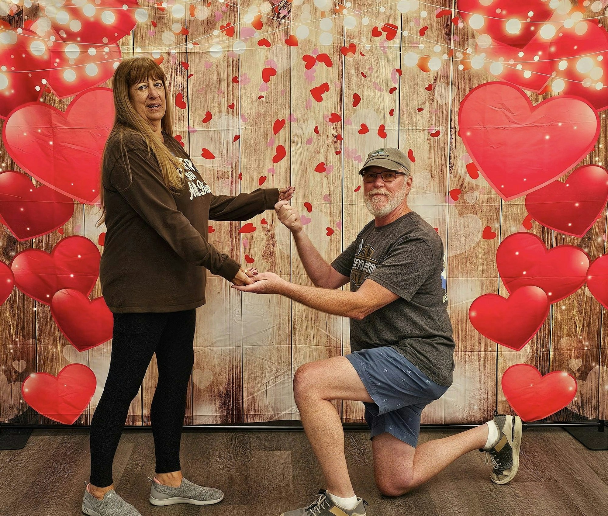 Two people posing with a heart-themed backdrop, one person is kneeling and holding the other's hand. #romantic #valentines