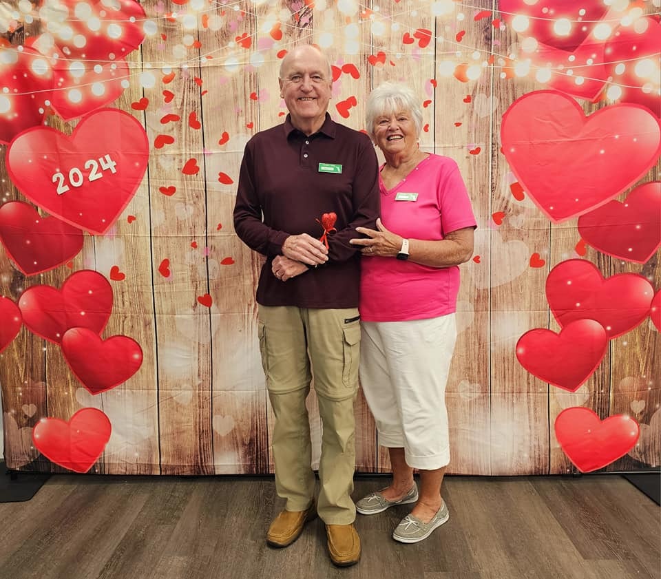 A smiling couple posing in front of a Valentine's Day themed backdrop with hearts and "2024" written on it.