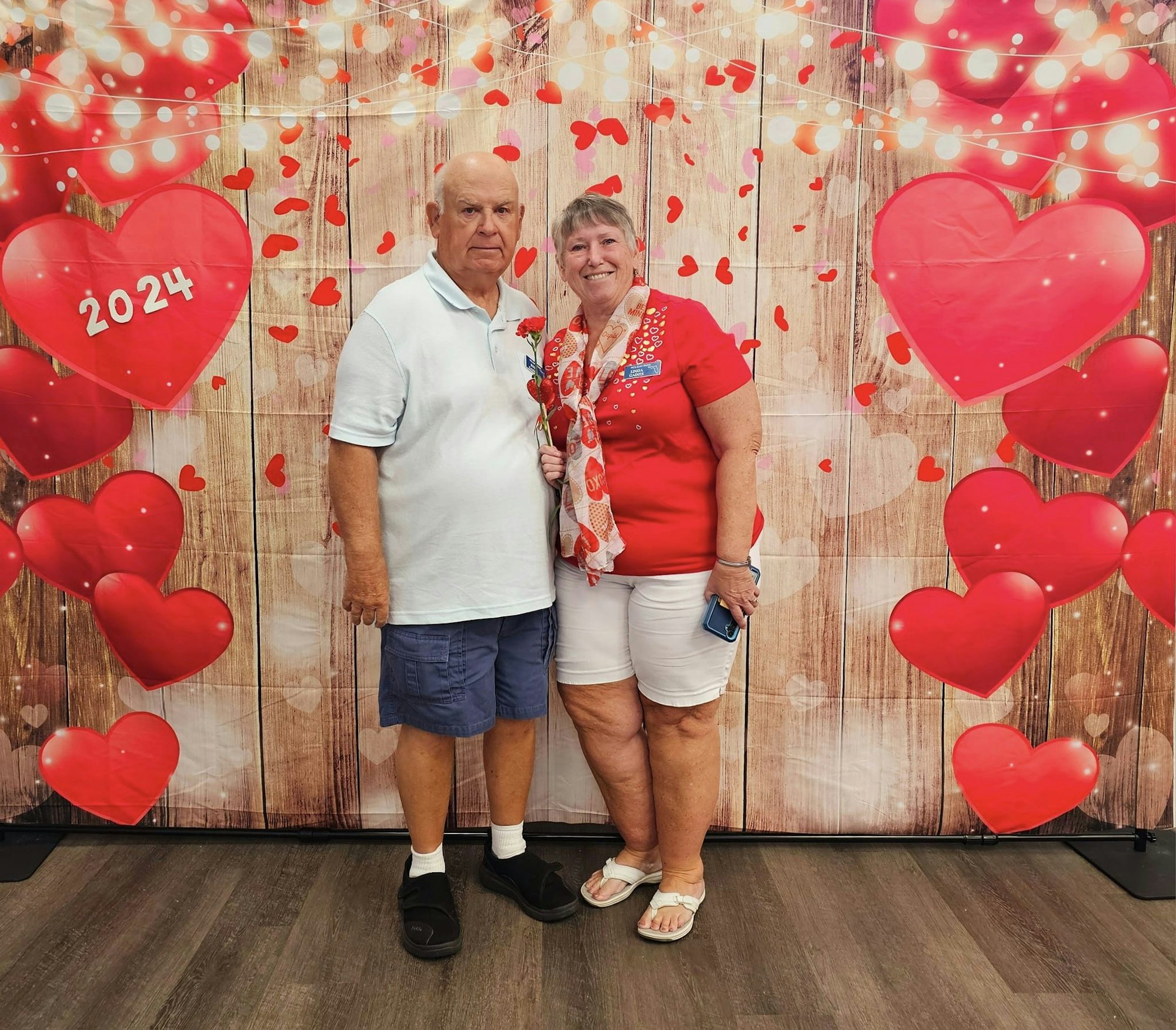 A couple posing in front of a Valentine's themed background with hearts and "2024" written on one heart.
