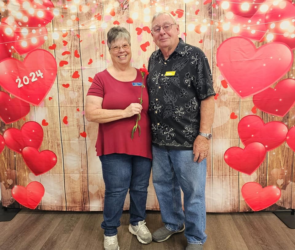 A couple posing with a rose against a romantic-themed backdrop with hearts and lights.