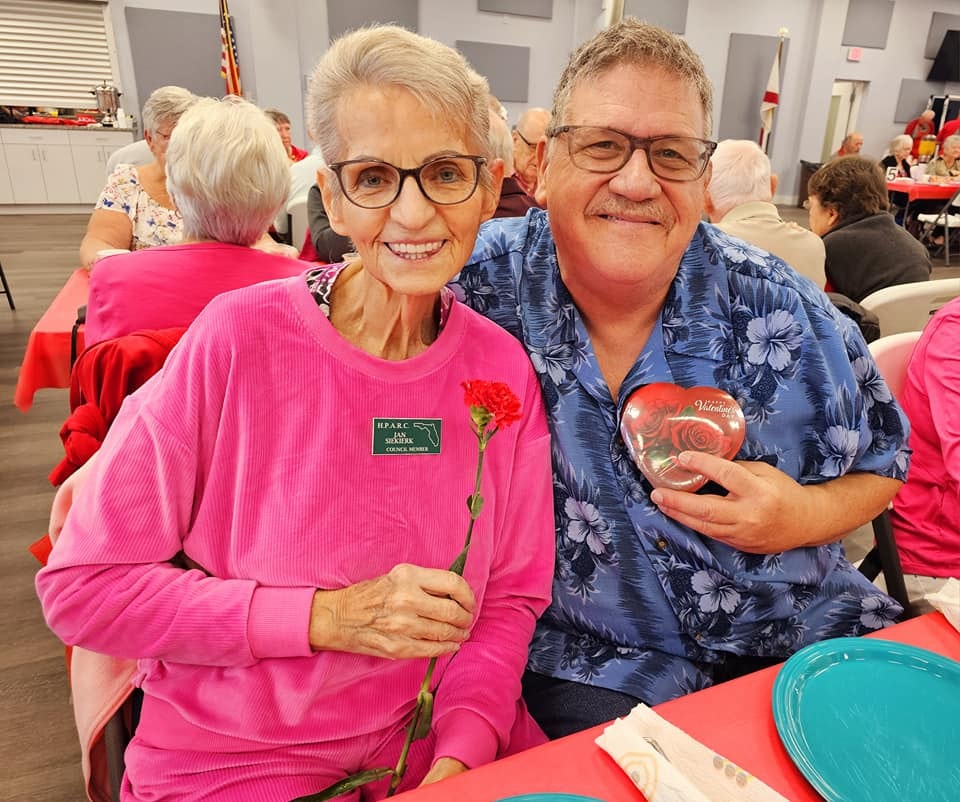 A smiling woman in pink holds a flower next to a man in a blue shirt, both celebrating at a festive gathering.