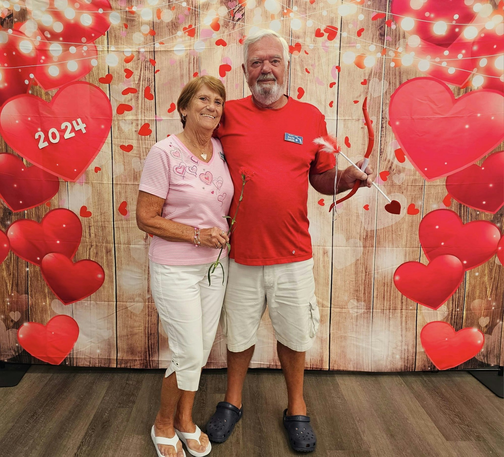 An older couple posing together with a Valentine's Day theme of hearts and "2024" sign.