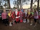 A group of people dressed in festive costumes, including Santa, gathered outdoors among trees during a celebration.