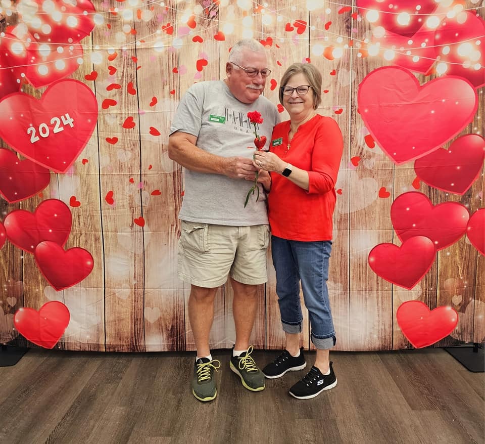 A couple holding a flower with a romantic heart-themed backdrop.