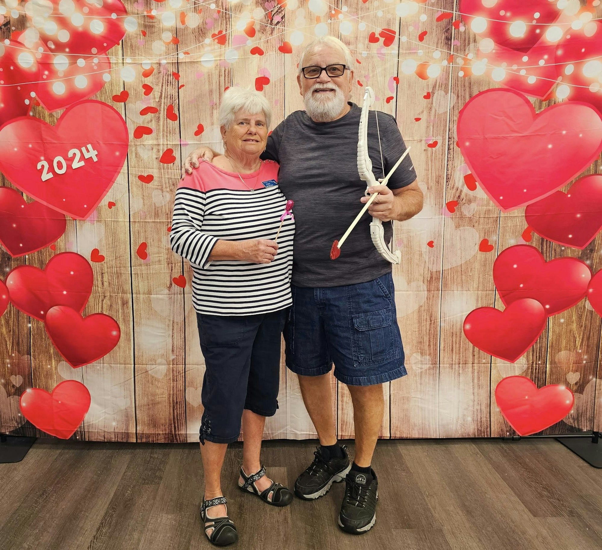 A couple stands before a heart-themed backdrop with "2024" on it, holding toy arrows, and smiling.