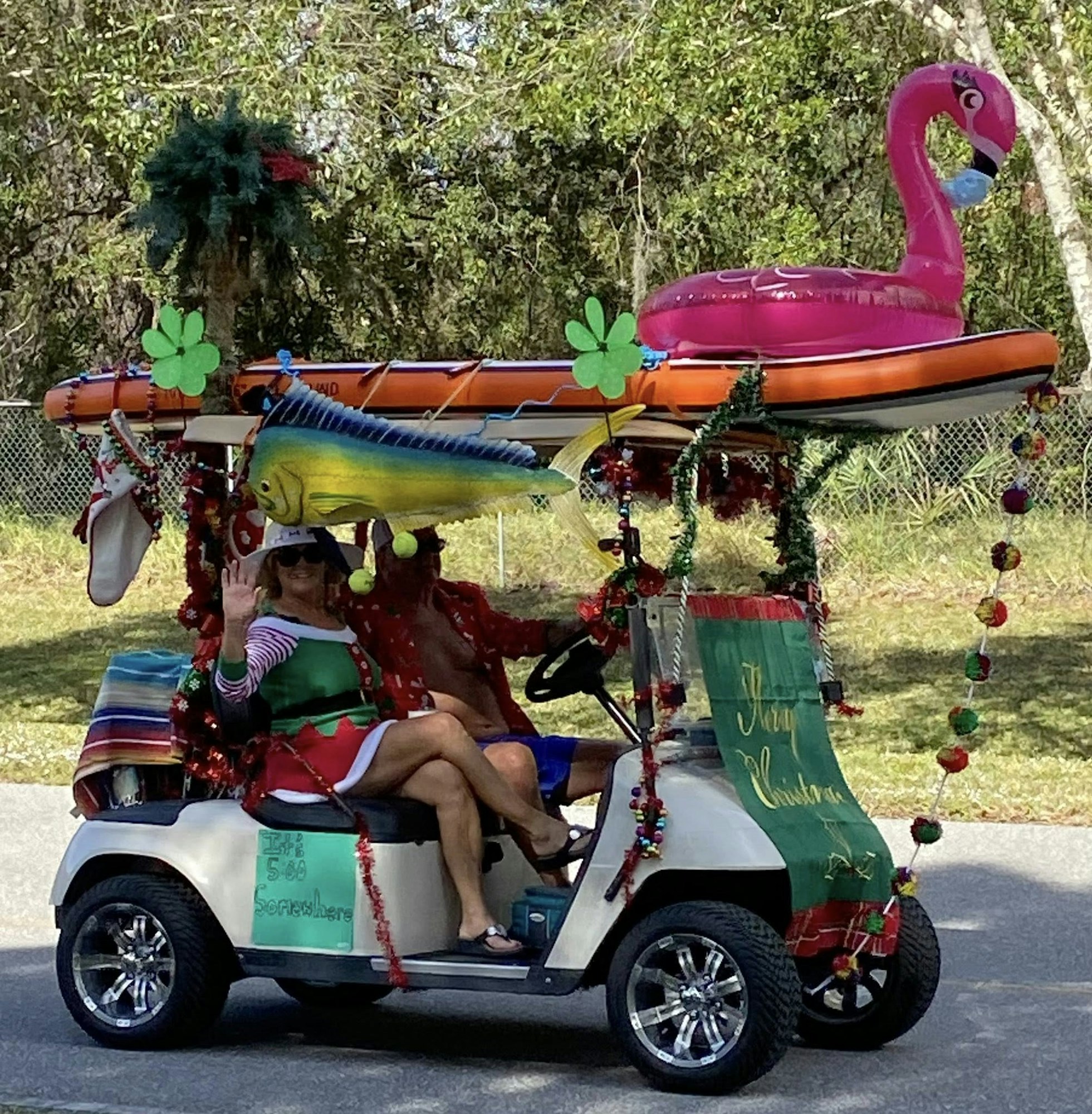 A decorated golf cart features festive items like a pink flamingo, surfboard, and colorful ornaments, with two people inside.