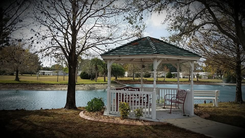 A serene gazebo by a pond, surrounded by trees, with benches and a peaceful landscape in the background.