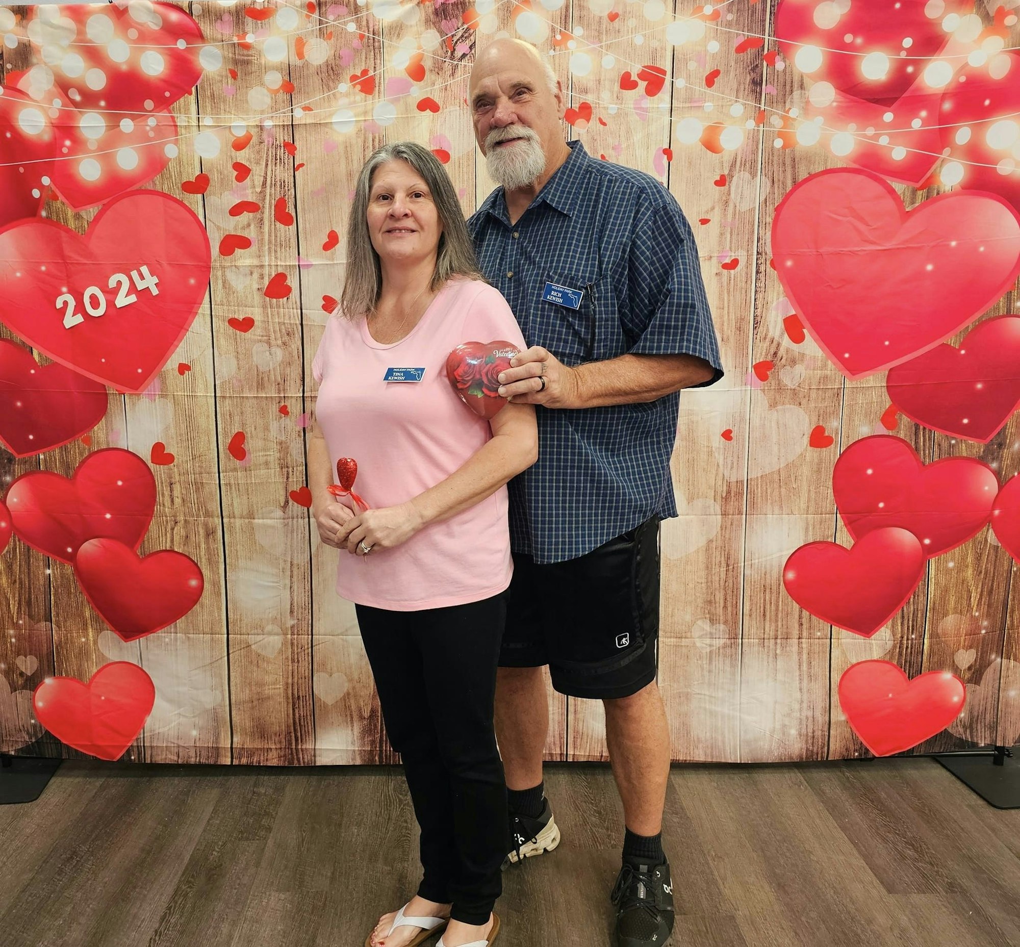 Two people posing with heart decorations and a "2024" sign, in a Valentine's Day-themed setup.