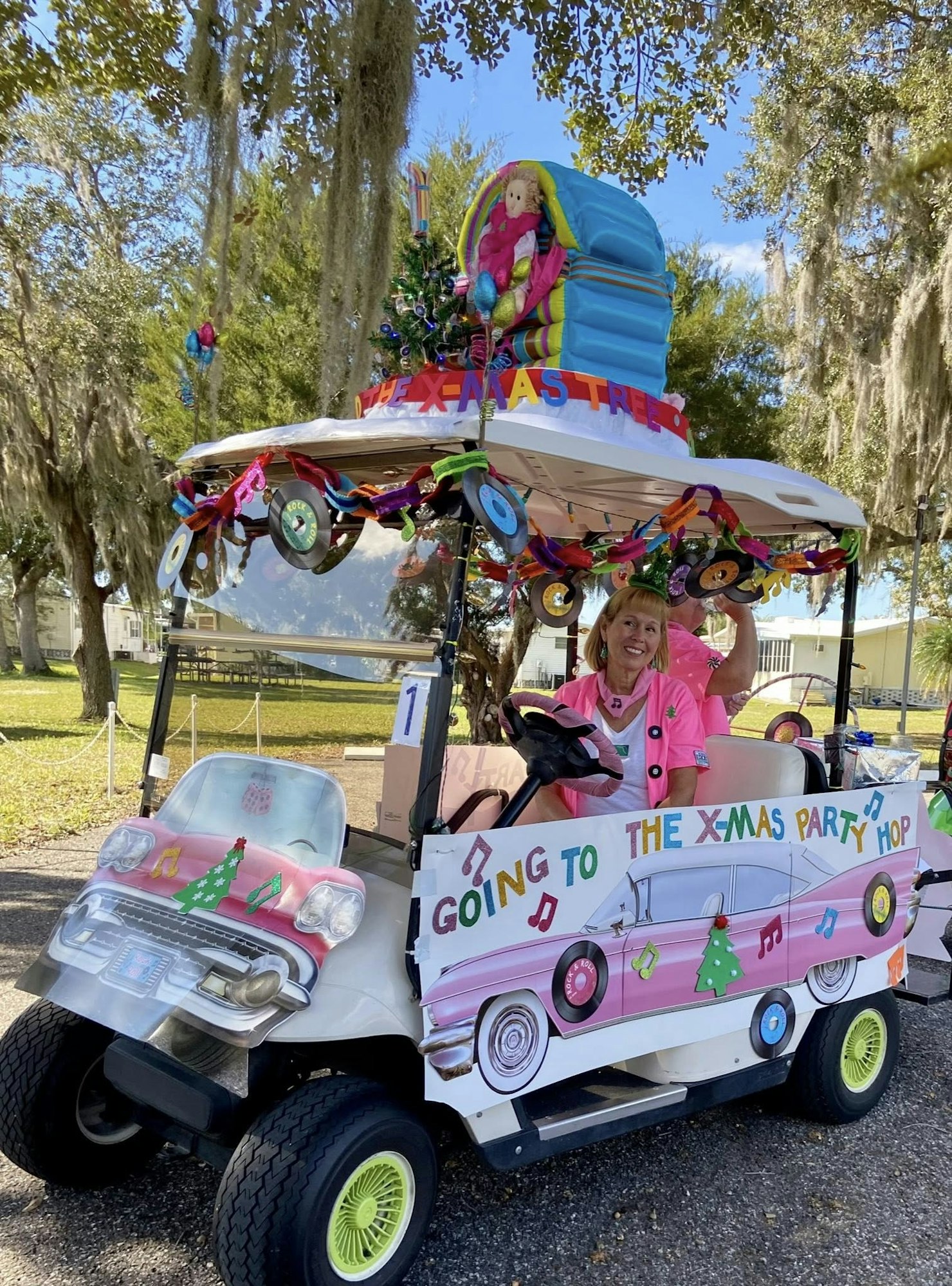 A festive golf cart, decorated for Christmas, featuring a cheerful driver in a pink outfit, heading to a holiday party.