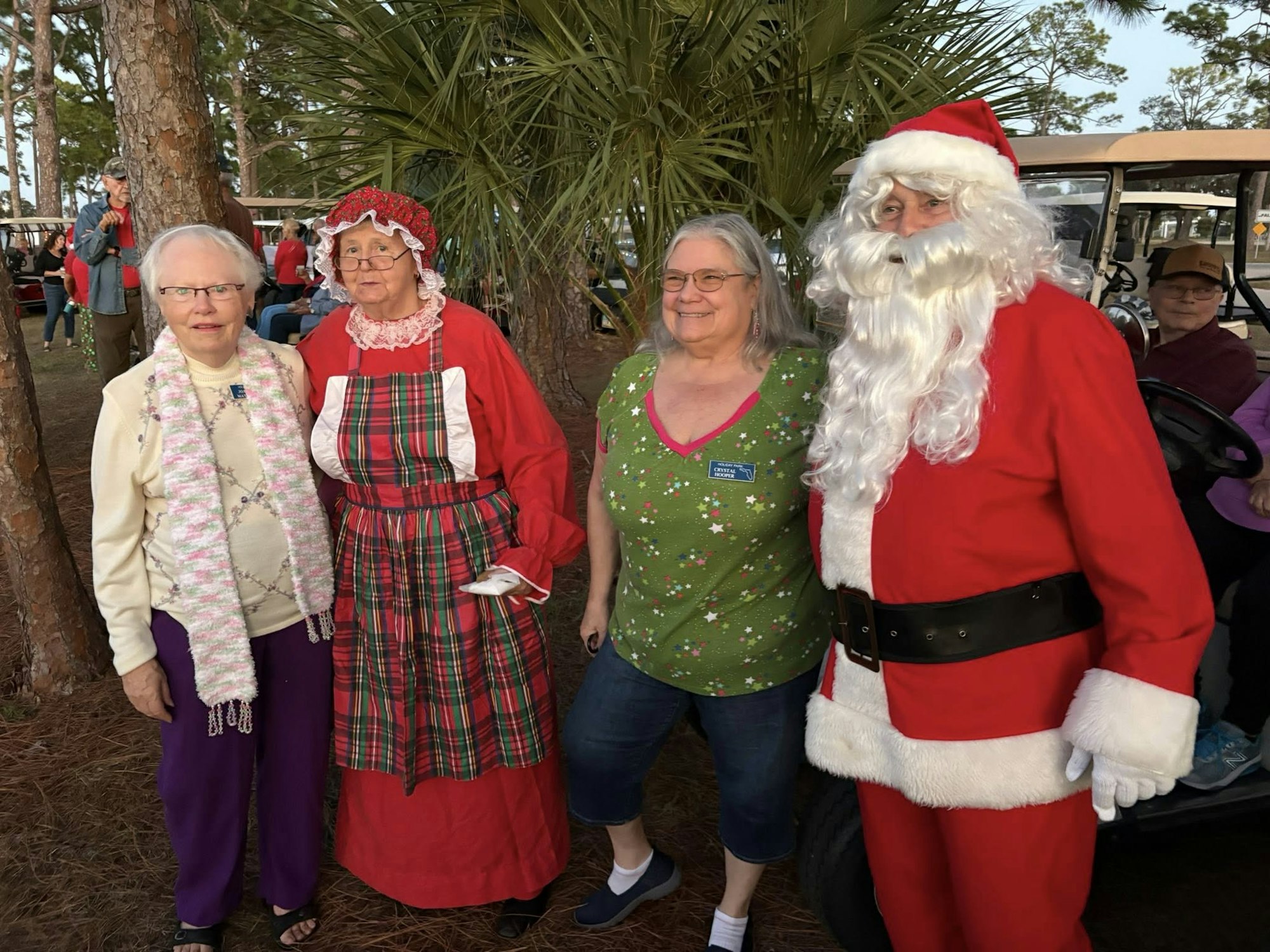 A festive gathering with three women and Santa Claus, all dressed in holiday attire, set in a park-like environment.