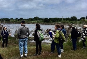 Group of people outdoors, possibly on a guided tour, near a body of water with grass and cloudy skies.