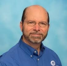 The image shows a man with glasses and a bald head, wearing a blue shirt, set against a light blue background.